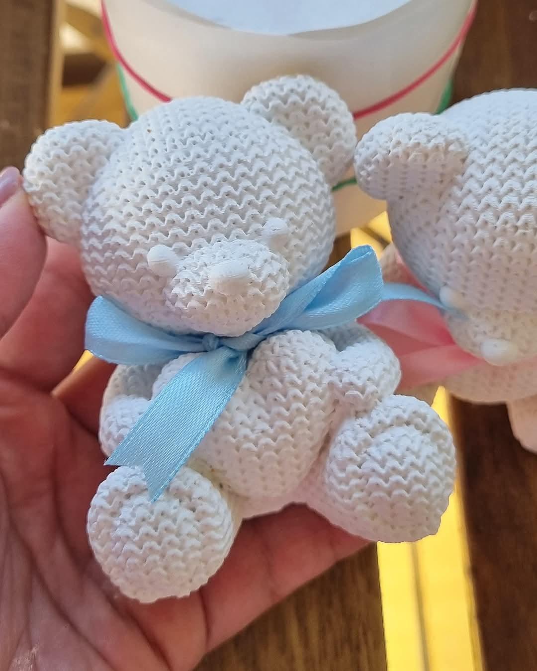 Ursinho de croché branco com fita azul, em posição frontal com dois exemplares