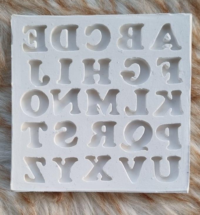 Molde de alfabeto quadrado com cavidades de letras em relevo, cor clara.