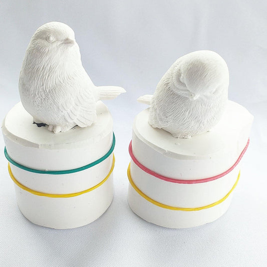 Par de moldes 3D de passarinho branco em bases cilíndricas com fitas coloridas